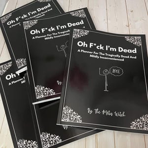 May include: Several black planners are stacked, each with the title "Oh F*ck I'm Dead" in white. The cover text reads "A Planner For The Tragically Dead And Mildly Inconvenienced", with a stick figure and a "BYE" speech bubble.