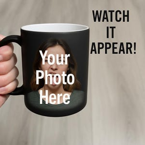 May include: Black ceramic mug with a white interior, featuring a photo of a person and the text "Your Photo Here" and "WATCH IT APPEAR!". The mug is held by a hand.