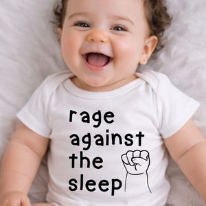 May include: A smiling baby wearing a white onesie with the text "rage against the sleep" and a fist graphic. The baby has curly brown hair and is lying on a white surface.