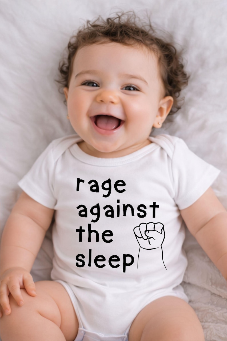 May include: A smiling baby wearing a white onesie with the text "rage against the sleep" and a fist graphic. The baby has curly brown hair and is lying on a white surface.
