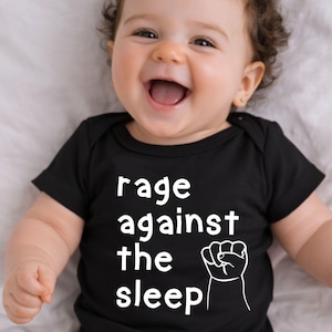 May include: A smiling baby wearing a black onesie with the words "rage against the sleep" and a white fist graphic. The baby has curly brown hair and is lying on a white surface.