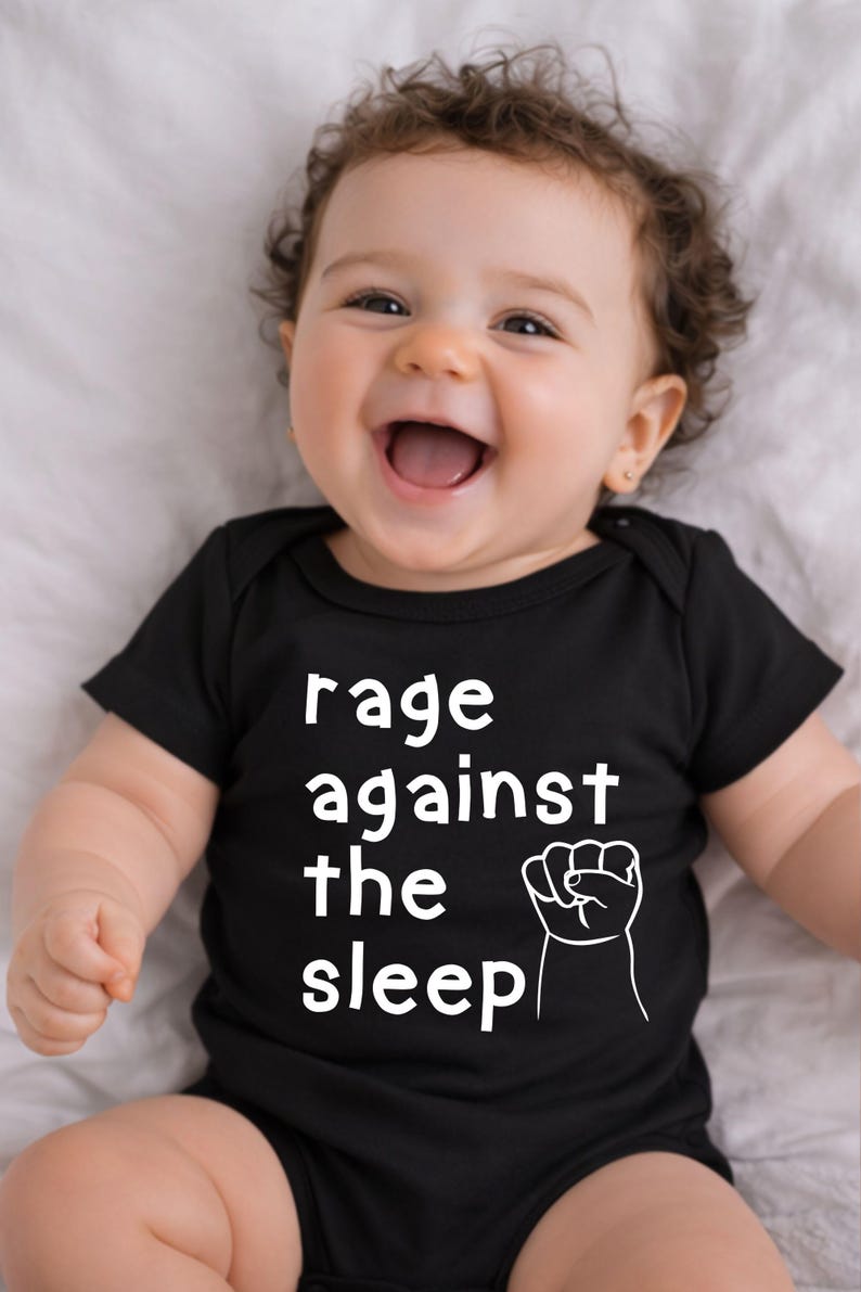 May include: A smiling baby wearing a black onesie with the words "rage against the sleep" and a white fist graphic. The baby has curly brown hair and is lying on a white surface.