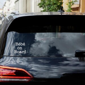 Op de afbeelding: Een zwarte auto met een witte sticker op de achterruit met de tekst "Bébé aan boord".