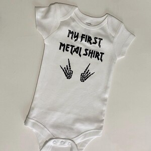 Może przedstawiać: Biały śpioszek dla niemowlęcia z czarnym napisem "My First Metal Shirt" i dwoma symbolami szkieletowych dłoni.
