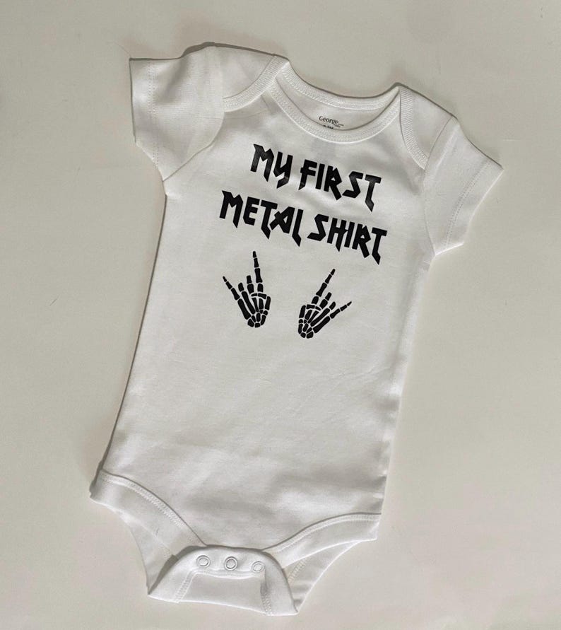 Może przedstawiać: Biały śpioszek dla niemowlęcia z czarnym napisem "My First Metal Shirt" i dwoma symbolami szkieletowych dłoni.