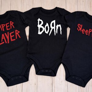 May include: Three black baby onesies with different red and white text. The onesie on the left says "Diaper Slayer", the middle one says "Born", and the right one says "Sleep Not". The onesies are displayed on a light wood surface.