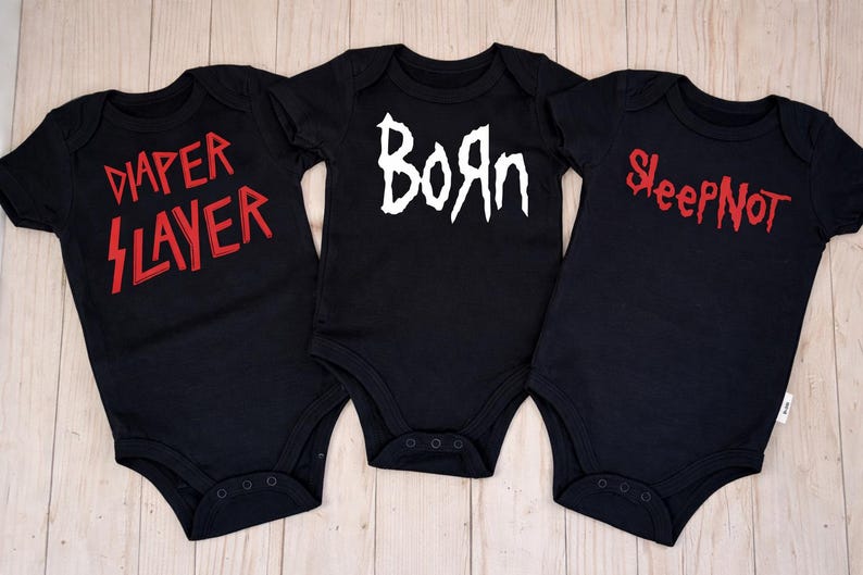 May include: Three black baby onesies with different red and white text. The onesie on the left says "Diaper Slayer", the middle one says "Born", and the right one says "Sleep Not". The onesies are displayed on a light wood surface.