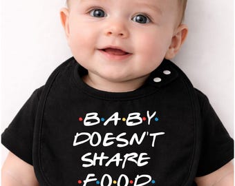 May include: A smiling baby wearing a black bib and t-shirt. The bib has the text "BABY DOESN'T SHARE FOOD" in white, with the word "BABY" in red, blue, and yellow dots. The baby has light skin and blue eyes.