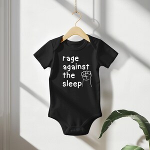 May include: Black baby bodysuit with the text "rage against the sleep" in white, along with a white fist graphic. The bodysuit is hanging on a wooden hanger against a white background.