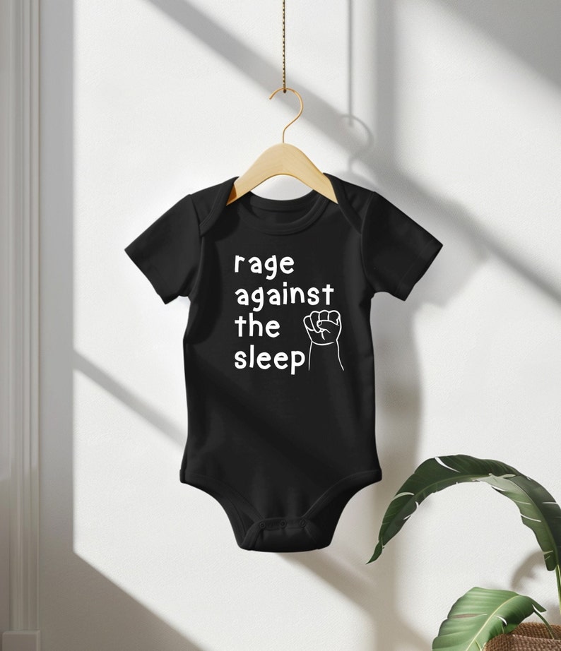 May include: Black baby bodysuit with the text "rage against the sleep" in white, along with a white fist graphic. The bodysuit is hanging on a wooden hanger against a white background.