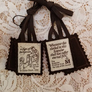 May include: Two brown scapulars with dark brown cords. Each scapular has a rectangular patch with a decorative border. One patch features an illustration and text: "a Sign of Salvation, Our Lady of Mt. Carmel to St. Simon Stock - 1251". The other patch has text: "Whosoever dies clothed in this Scapular shall not suffer eternal fire".