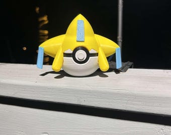 May include: A yellow and white 3D-printed figure, resembling a stylized star with blue accents. The figure has a round, white base with a black and white circle in the center. It is resting on a white painted surface.