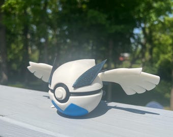 May include: A white and blue spherical figurine with stylized wings. The figurine has a blue and black accent design and a circular detail on the front. The wings are white and extend outward. The object is set against a blurred green background.