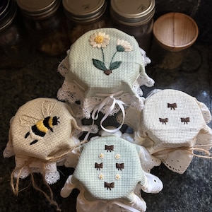 Puede incluir: Cuatro tapas de tarros con diseños bordados. Una presenta un abejorro, otra margaritas y dos con patrones de lazo. Cada tapa es de color claro con ribete de encaje blanco y atada con hilo o cinta. Los tarros son visibles en el fondo.