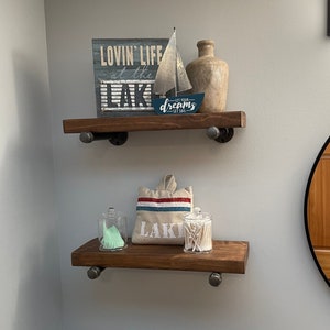 May include: Two wooden shelves with black metal brackets. The top shelf has a sign that says "Lovin' Life at the Lake" with a small sailboat and a brown ceramic vase. The bottom shelf has a white canvas bag with the word "Lake" on it, a clear glass jar with cotton swabs, and a clear glass bottle with a green liquid.