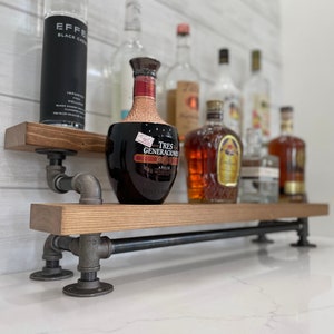 May include: A two-tiered wooden shelf with black metal pipe supports. The shelf is filled with various bottles of liquor, including a bottle of Tres Generaciones Anejo tequila.