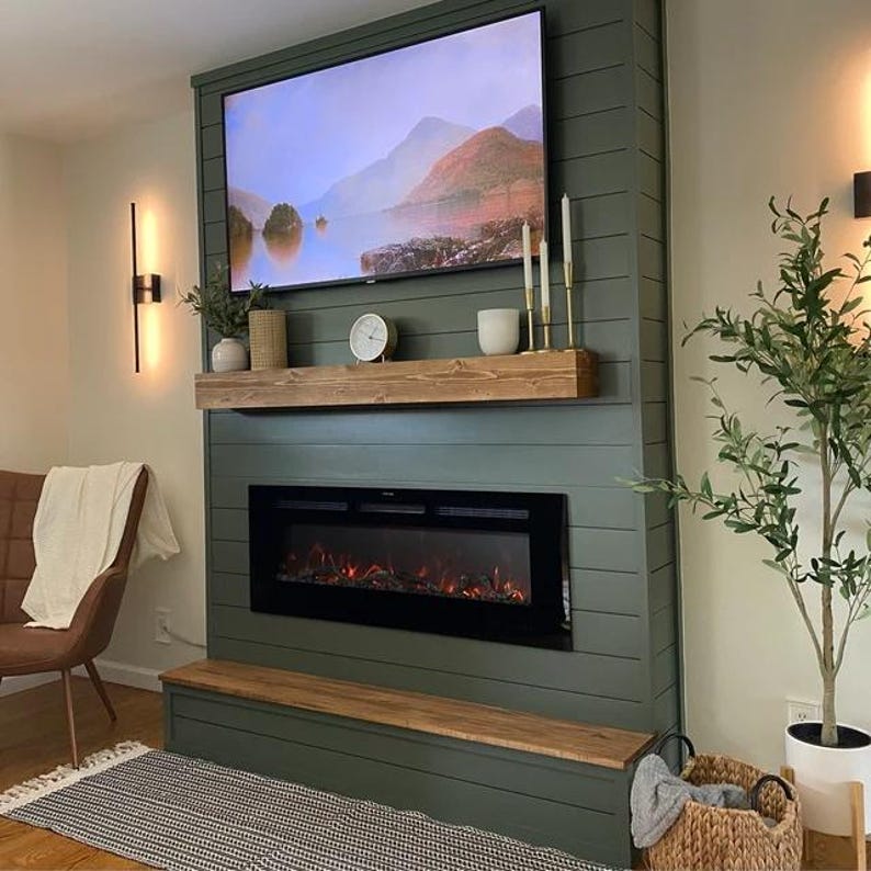May include: A modern living room entertainment center with a large flat-screen TV mounted above an electric fireplace. The wall is painted a dark green and features a wooden mantel with decorative candles and a clock. A bench and rug complete the cozy space.