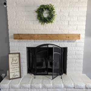 Op de afbeelding: Een witte bakstenen open haard met een houten schouw en een zwart metalen haardrooster. Een bord met de tekst "Today I marry my Best Friend the one I laugh with, live for, dream with. Love." staat aan de linkerkant van de open haard. Een groene krans hangt boven de schouw.