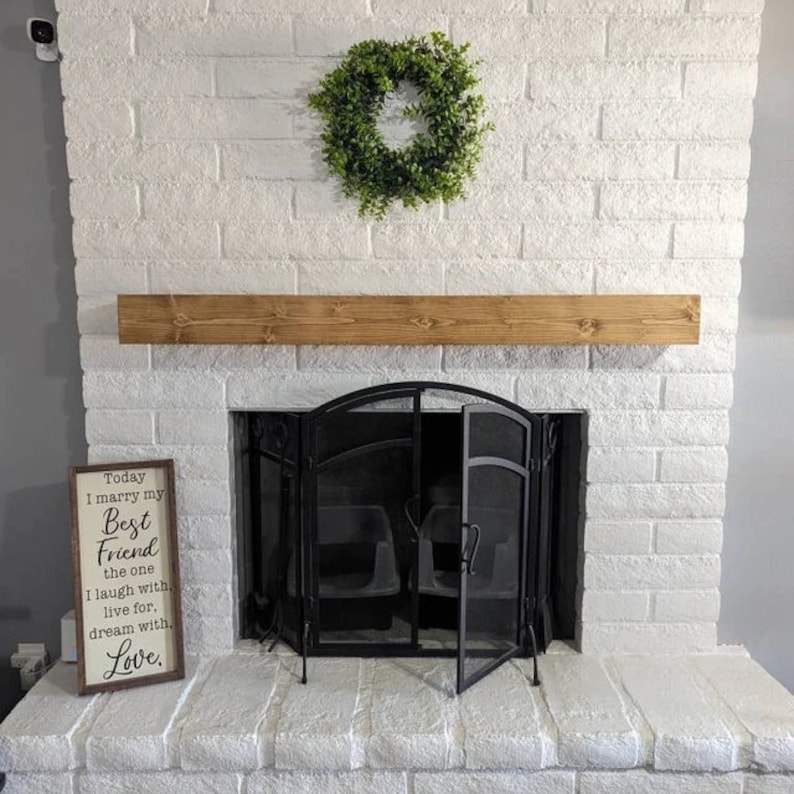 Op de afbeelding: Een witte bakstenen open haard met een houten schouw en een zwart metalen haardrooster. Een bord met de tekst "Today I marry my Best Friend the one I laugh with, live for, dream with. Love." staat aan de linkerkant van de open haard. Een groene krans hangt boven de schouw.