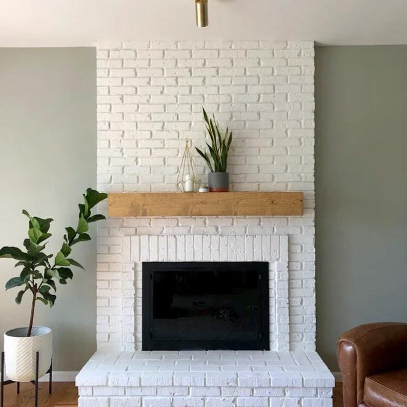 May include: A white brick fireplace with a black firebox and a wooden mantel. A potted plant sits on the mantel, and another plant is in a white pot on the floor. The wall is painted a light green.