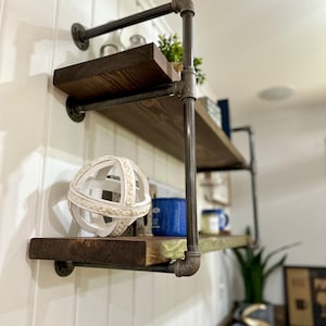 May include: Two tiered wooden shelves with black metal pipe supports. The shelves are decorated with a white decorative sphere, a blue mug, and a green plant.