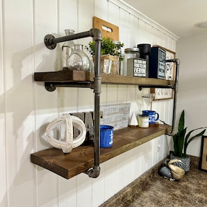 May include: Two-tiered wooden shelf unit with black metal pipe supports. The shelves are decorated with various items, including a white globe, a blue mug, a glass jar, and a small potted plant. The shelf is mounted on a white wall.