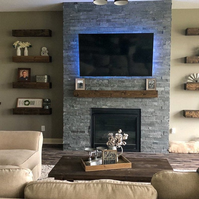 May include: A living room with a stone fireplace, a large TV, and wooden floating shelves. A wooden coffee table holds a tray with decorative items. The room has a neutral color palette with a cozy, inviting atmosphere.