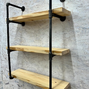 May include: Three-tiered black pipe shelf unit with wooden shelves. The shelves are made of natural wood and are supported by black metal pipes. The unit is mounted to a wall.