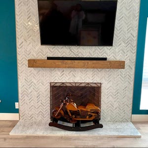 May include: A wooden rocking motorcycle in shades of brown and tan sits in front of a fireplace. The fireplace has a decorative screen, a wooden mantel, and a herringbone tile design. A large TV is mounted above the mantel.