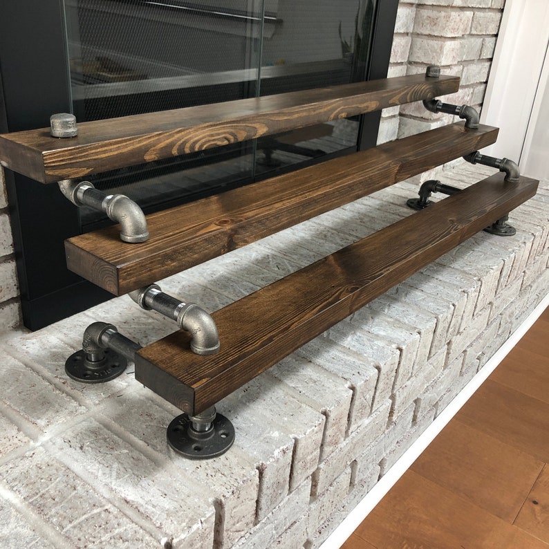Puede incluir: Una repisa de chimenea de tres niveles hecha de madera marr&oacute;n oscuro con soportes de tuber&iacute;a de metal negro. La repisa est&aacute; montada en una pared de ladrillo blanco.