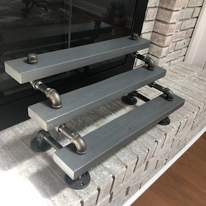 Puede incluir: Estanter&iacute;a de tres niveles hecha de tablones de madera gris oscuro y tuber&iacute;as de metal negro. La estanter&iacute;a est&aacute; montada en una pared de ladrillo blanco.