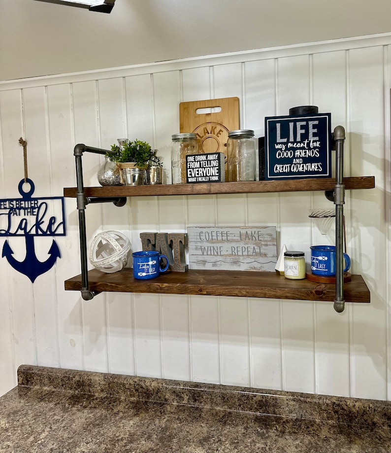 May include: Two wooden shelves with black metal pipe supports. The shelves are decorated with various items, including a blue anchor, a sign that says "Life was meant for good friends and great adventures", a sign that says "Coffee - Lake - Wine - Repeat", and a sign that says "One drink away from telling everyone what I really think".