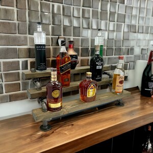 Puede incluir: Una estanter&iacute;a de madera con tuber&iacute;as de metal negro, que contiene varias botellas de licor. Las botellas incluyen vodka, whisky, ron y tequila. La estanter&iacute;a est&aacute; sobre una encimera de madera.