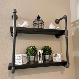 May include: Two-tiered black metal shelf with industrial pipe accents. The shelf is decorated with two potted plants, two white bird figurines, a birdcage, and three wooden signs that read "Hello", "Be Seated", and "Est. 2018".