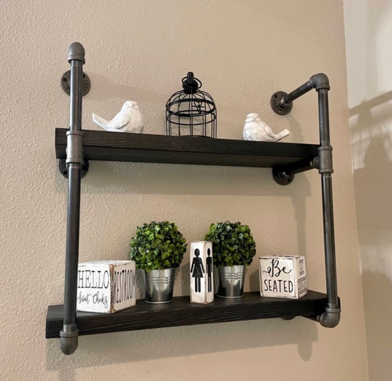 May include: Two-tiered black metal shelf with industrial pipe accents. The shelf is decorated with two potted plants, two white bird figurines, a birdcage, and three wooden signs that read "Hello", "Be Seated", and "Est. 2018".