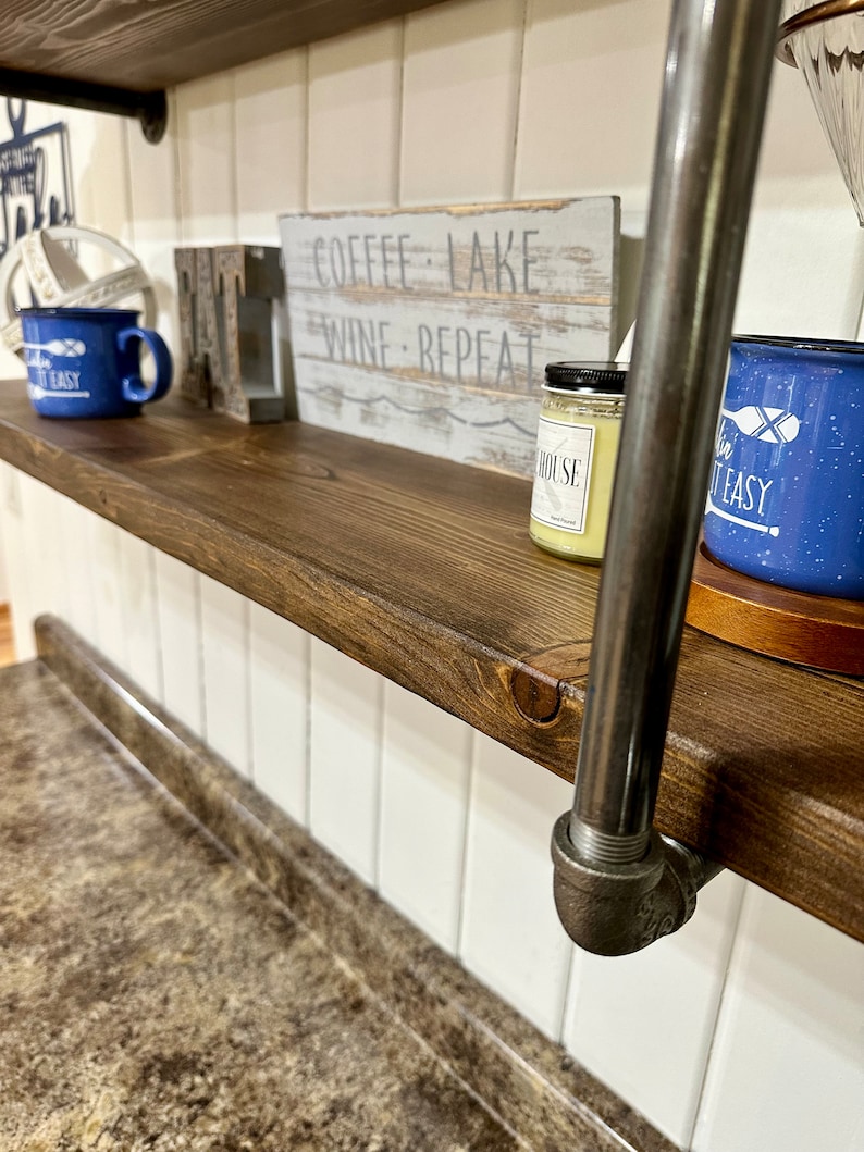 May include: A wooden shelf with a metal pipe support. The shelf is decorated with two blue mugs, a wooden sign that says "Coffee Lake Wine Repeat", and a candle. The shelf is mounted on a white tiled wall.