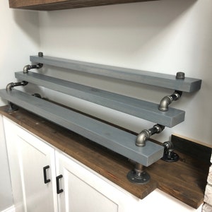 Puede incluir: Tres estantes de madera gris con soportes de tuber&iacute;a de metal negro. Los estantes est&aacute;n montados en una pared y est&aacute;n escalonados en altura.