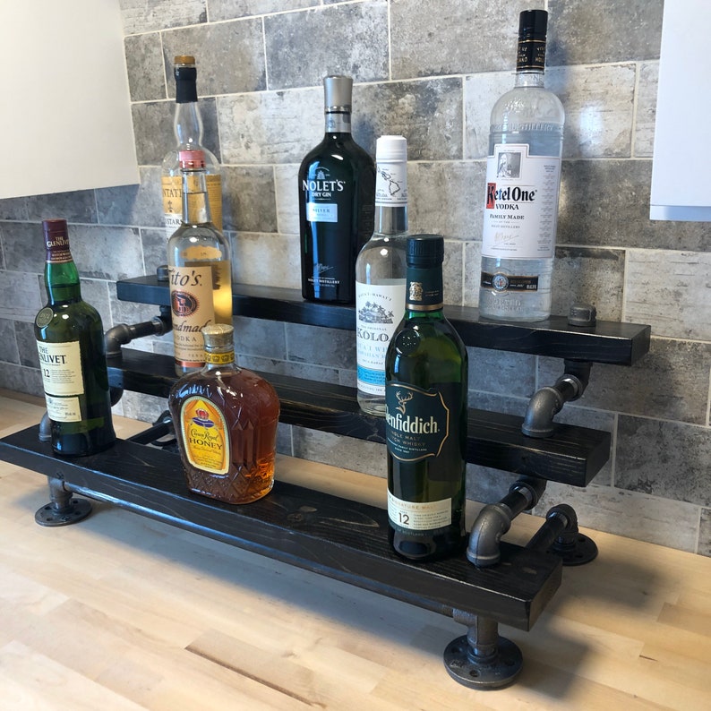 Puede incluir: Estanter&iacute;a de madera negra de tres niveles con soportes de tuber&iacute;a de metal negro. La estanter&iacute;a est&aacute; llena de varias botellas de licor, incluyendo Tito's Vodka, Crown Royal y Glenfiddich.