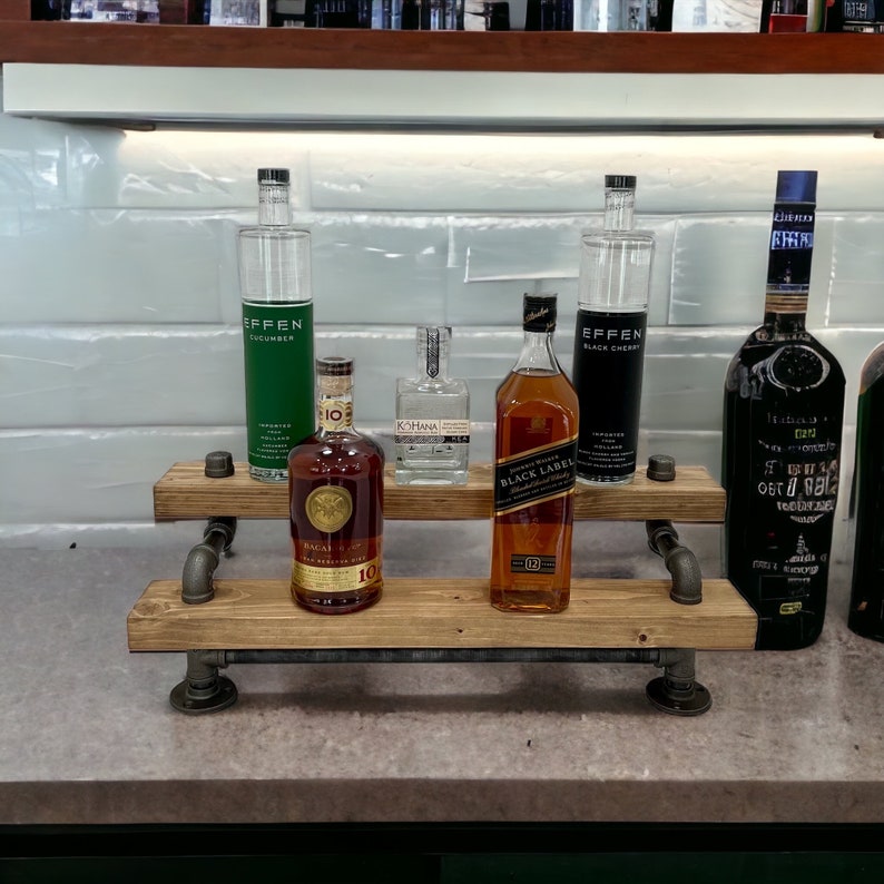 Puede incluir: Una estanter&iacute;a de madera de dos niveles con soportes de tuber&iacute;a de metal negro, que contiene una variedad de botellas de licor. Las botellas incluyen vodka Effen con pepino, vodka Effen con cereza negra, whisky escoc&eacute;s Johnnie Walker Black Label y una botella de ron. La estanter&iacute;a est&aacute; sobre una encimera gris.