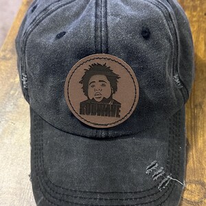 May include: A black distressed baseball cap with a brown leather patch featuring a black and white image of a person with the word "RODWAVE" printed in white.