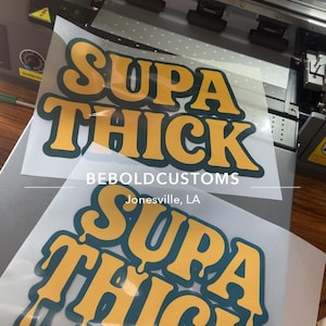 May include: Two clear sheets with the words "SUPA THICK" in yellow bubble letters with a teal outline. The text "BEBOLDCUSTOMS Jonesville, LA" is below the main text. The sheets are on a gray surface.