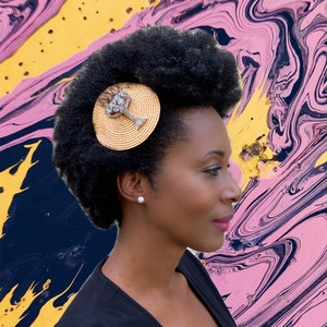 Puede incluir: Una mujer con cabello castaño oscuro lleva un accesorio para el cabello dorado y negro con un diseño de diamantes de imitación. El accesorio tiene la forma de un pequeño sombrero con ala.