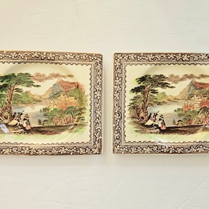 May include: Two square ceramic plates with a brown floral border. Each plate features a scenic landscape with a lake, mountains, and trees. The plates are in a vintage style.