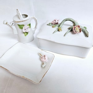 Puede incluir: Un juego de porcelana blanca con una pequeña regadera, una caja de alhajas rectangular con un diseño floral y un pequeño plato cuadrado con una sola rosa rosa.