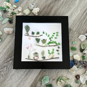 May include: A framed artwork featuring a collection of green and brown sea glass arranged to resemble potted plants. The artwork is set against a white background and includes a small pink flower arrangement. The frame is black and the artwork is signed "MH".