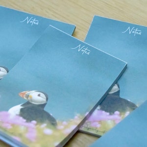 May include: A stack of notebooks with a blue cover featuring a puffin illustration. The word "Notes" is written in cursive at the top. The puffin is surrounded by pink flowers.