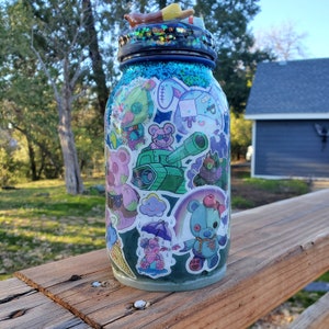 May include: A blue glass jar with a glittery finish decorated with colorful cartoon stickers. The stickers feature characters, a tank, and a cloud.
