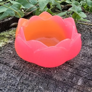 May include: A bowl shaped like a lotus flower with a smooth, translucent finish. The object features a vibrant gradient of colours, transitioning from a bright neon pink on the outer petals to a warm, glowing orange in the centre of the bowl.