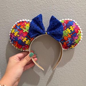 Puede incluir: Diadema con orejas de ratón con un patrón de rompecabezas colorido. Las orejas están adornadas con pompones blancos y un gran lazo azul con lentejuelas se encuentra entre ellas. La diadema es blanca.