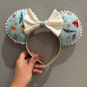 May include: A headband with two round, light blue ears featuring a colorful aquatic print with fish and coral. A large white bow sits between the ears. The headband is white with a gold interior.